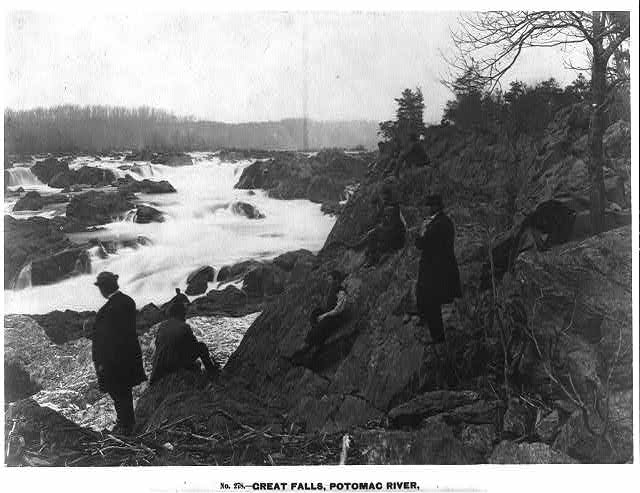 Great Falls of the Potomac River rushing through a rocky gorge, with several people standing on the rocks above the water.